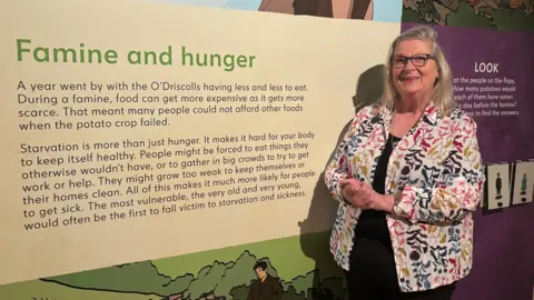 A woman stands in front of a display board in a museum. The display board is titled Famine and hunger. A woman in a brightly patterned shirt stands alongside it. She had blonde hair and is wearing glasses. Her hands are clasped in front of her and she is smiling. 