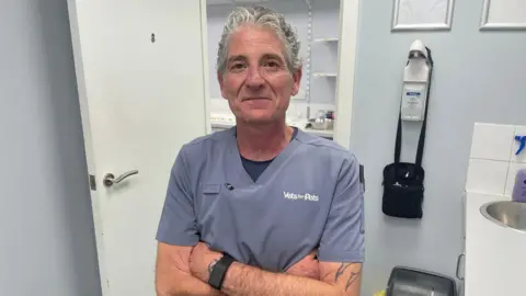 A man with grey curly hair stands with arms folded in a veterinary treatment room. He wears grey Vets for Pets scrubs and has a tattoo on one forearm. Beside him are a sink, a hand sanitiser dispenser and a yellow clinical waste bag.