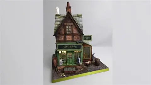 Nikki Lowe A cake designed like a shop. It has a pointed roof with a white owl on the left hand side. There is a sign on the front of the shop.