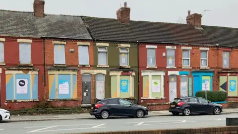 A wider view of the row of terraced houses with their windows and doors shuttered with metal sheets.