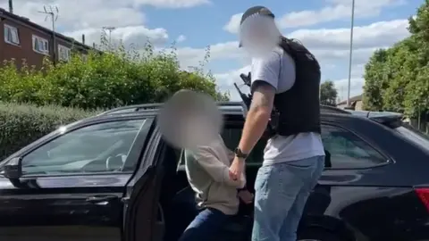 A plain-clothes police officer guides an arrested suspect into the back of a car. Both men's faces are blurred