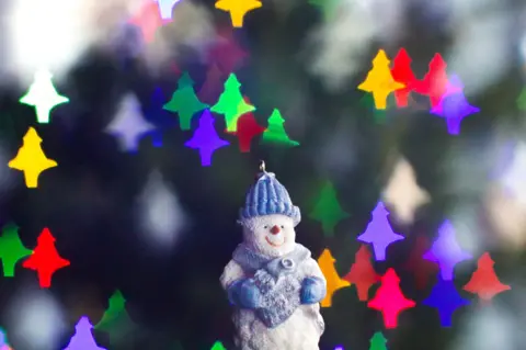 Miles Harrison Close-up of snowman ornament with twinkling Christmas lights in the background in the shape of Christmas trees.