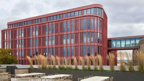 The exterior of the Forum building in Gloucester. The building has a red facade and chairs and tables outside.