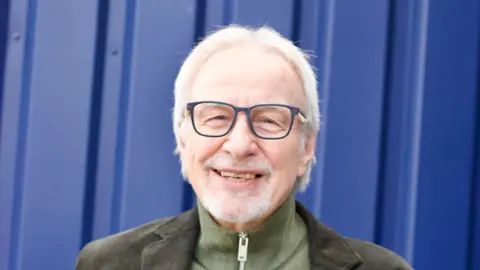 Gatehouse A head and shoulders image of a man wearing glasses and a dark green jacket over a lighter green top with a zip on it. He is smiling and standing in front of what looks like a blue metal container or door. 