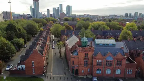 An old brick building can be seen from above surrounded by brick houses, trees and sky scrapers in the distance. 