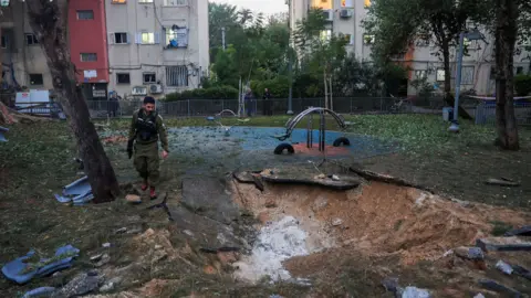 Reuters A man in a khaki uniform stands in a playground in front of a block of flats, looking down at a crater in the ground