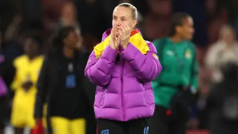 PA Media A woman with tied back blonde hair and wearing a purple and yellow padded coat and England shorts is clapping. She is standing in a football ground.