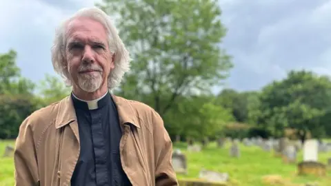 BBC The Reverend Tim Heaney, wearing a clerical collar and a brown coat, stands in a grassy graveyard with scattered headstones, trees and hedges in the background under an overcast sky.