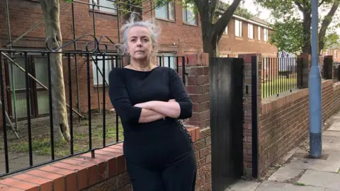 Thelma Clark has grey hair and a black top and black trousers. She is standing with her arms crossed in front of a brick wall. 