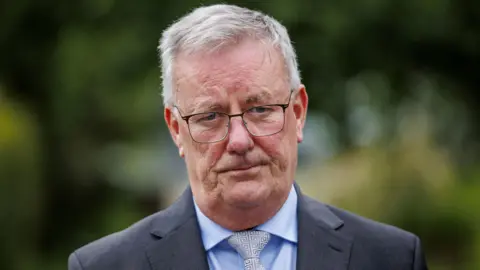Mike Nesbitt is wearing a grey blazer, blue shirt and grey tie. He has black glasses on his face. He has short grey hair. The background is blurry. 