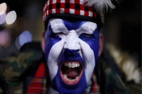 Reuters Close up of a Scotland fan with a saltire painted on his face. He is baring his teeth in celebration and his eyes are closed. He is wearing a red tartan hat and scarf.