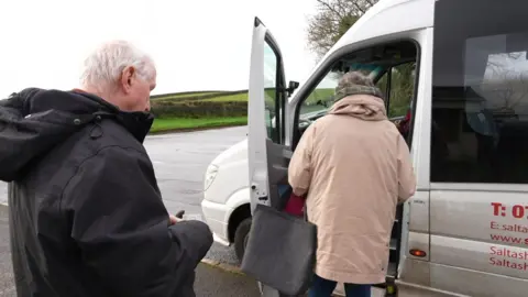 A man and a woman are queuing up to step into the white community bus which has its door open. There are hills beyond the road. The man is holding a bus pass and the lady has a shopping bag hanging over her arm. The bus has contact details printed in red on the side.