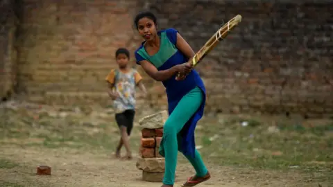 A woman, wearing a blue and green outfit, swings a cricket bat on February 27, 2024 in Ranchi, India. Behind her is a makeshift wicket made of bricks and stones. A young boy wearing a T-shirt and shorts is visible in the background.