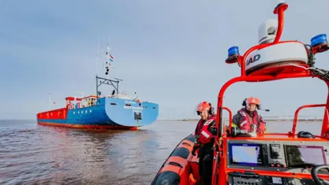 Humber Rescue On the right, a Humber Rescue lifeboat can be seen on the estuary with two men operating it and wearing red lifejackets and helmets. A large blue and red cargo vessel can be seen in front and to the left.