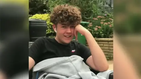 Stephens family Olly Stephens, a boy with dark curly hair and wearing a black -shirt. He's resting his head on one hand and smiling at the camera.