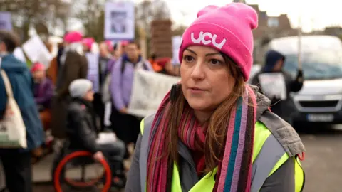 A woman with brown hair, wearing a yellow tabard and a pink hat with the letters UCU stands on a picket line