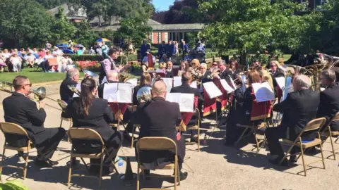A full brass band are playing for people lounging in a field. There are about two dozen people wearing black suits sitting in a semi-circle around the conductor. There are playing instruments like trumpets and French horns, while looking at sheet music on stands in front of them.