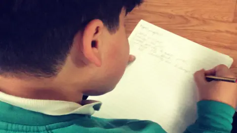 Sherine Lee Satti We are looking over the shoulder of a boy with dark hair and wearing a greenish blue jumper. He is writing on a white, ruled piece of paper.