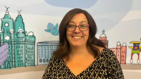 BBC/Claire Hamilton A woman with long brown hair and glasses stands, wearing a v-neck top stands in front of a giant poster featuring a brightly coloured cartoon of Liverpool's skyline. 