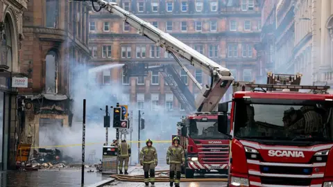 Fire fighters and fire engines at the scene of a major fire in Glasgow city centre - it is daytime and smoke from the blaze billows around the bottom of a building near Central Station