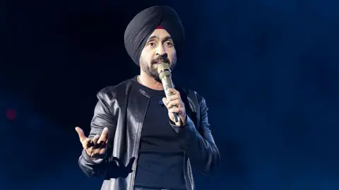 Diljit Dosanjh kicks off his Aura World Tour at BC Place on April 23, 2026, in Vancouver, British Columbia, Canada. He is wearing a black jacket over a black t-shirt and sporting a black turban, as he speaks on a microphone from the stage.