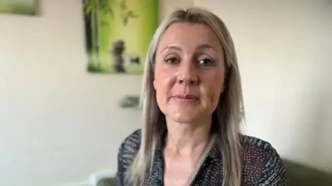 Emma Baugh/BBC Mandy Geraghty looks directly at the camera with a neutral expression. She has long blonde hair and is wearing a dark patterned top, and sits in front of a calm green picture.