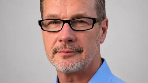 Close-up man with greying hair and greying beard, wearing glasses and a blue shirt