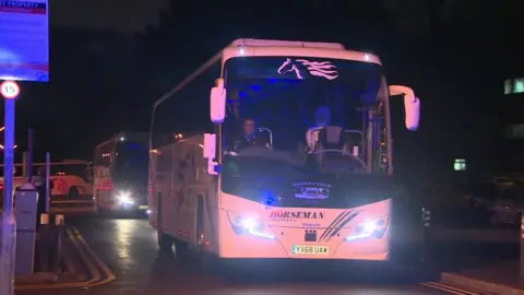 A white coach pulls into a hospital at night time. The coach driver can be seen not wearing a mask, while someone is stood next to him wearing face coverings and a body suit