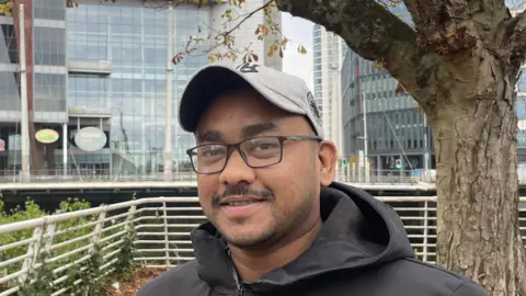 Rijvi Chowdhury smiling at the camera, he is wearing a grey cap and a black jacket. In front of the bridge at Wood Street at the Fitzhamon Embankment. 