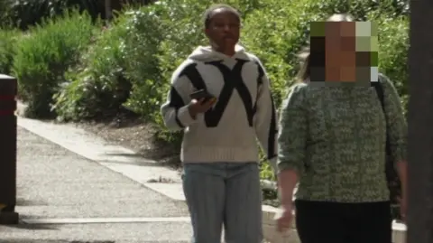 Elizabeth Adeagbo, who is black, has short hair and is wearing a white woollen jumper with a black pattern and jeans, walks along a pedestrian paved area holding a mobile phone in her right hand. 