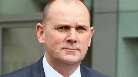 PA Media Head-and-shoulders image of Jim Gamble in a suit, looking directly at the camera with a neutral expression, taken outside a modern building.