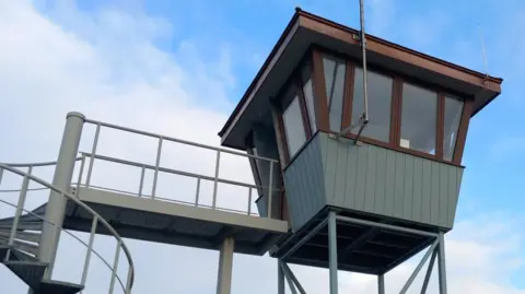 A grey coloured control tower. It stands on four legs and is accessed by a spiral staircase and connecting walkway