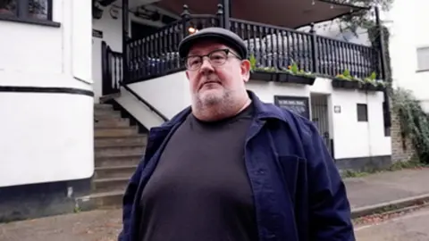 Handout Johnny Vegas stands outside the pub wearing a dark jumper and coat, flat cap and glasses