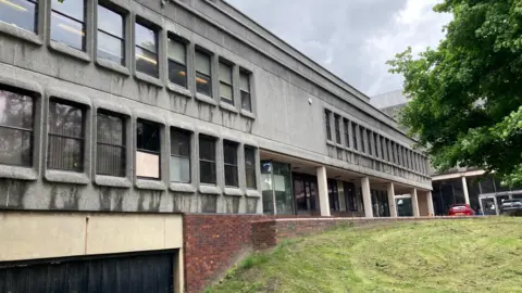 LDRS A grey 1960s brutalist building, situated next to a grassy patch of land. 