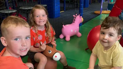 Three children play on inflatable animal hoppers in a soft play area at an indoor adventure playground