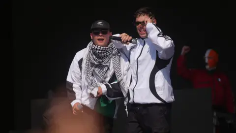 PA Media Two band members stand side by side on stage. They are both wearing white jackets and each hold a microphone. One of them has their elbow resting on his bandmate's shoulder as he appears to lift his sunglasses up. In the background, the third member can be seen holding his fist in the air. He is wearing a red jacket and a balaclava in the colours of the Irish flag.