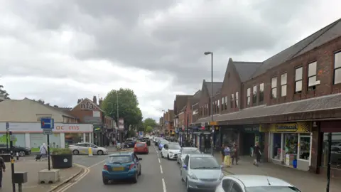 Google A high street busy with cars, with a road junction to the left. An acorns shop can be seen to the left, with a range of shop fronts on the right. Buildings on both sides are modern two to three storey structures built with dark red/brown brick. Pedestrians can be seen on both sides of the pavement.