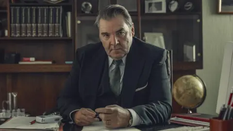 Ben Blackall/Netflix Brendan Coyle as Roy in Toxic Town. He is sitting at a desk with a bookcase behind him. He is wearing a black suit.