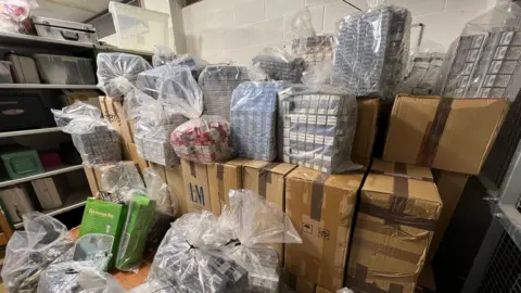 A very large pile of cardboard boxes and large plastic bags containing thousands of boxes of cigarettes inside a lock-up. There are shelves with containers on in the background.