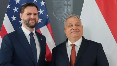 JD Vance and Orban stand together smiling, in front of their national flags