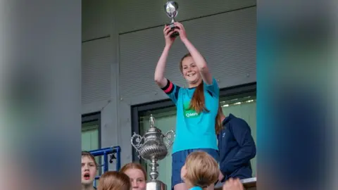 Clara has long brown hair in a ponytail. She is wearing a light blue football shirt. There is a large trophy beside her. There are children in the foreground.