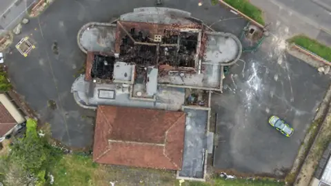 Ian Bateman A full aerial shot of a pub that has been severely damaged by fire. A police car is to the right. There is an area of grass and a large car park around the building. 