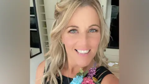 Kerri Deeley A woman with blonde hair smiles at the camera. She is wearing a black top with a corsage pinned to it. The corsage has blue, pink and purple flowers in it.