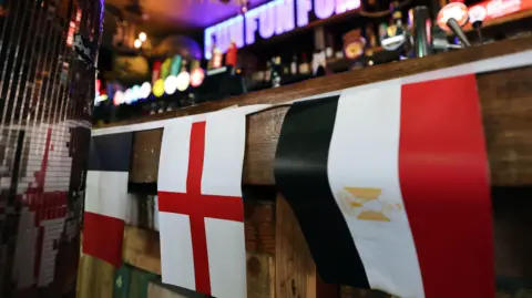 Flags on a banner across the front of a bar in a pub. At the centre is a white and red England flag. Behind the bar are lights and draughts. 