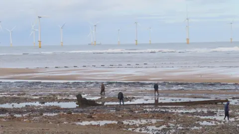 Weather Watchers / Sue J A beach at low tide. There are stones and pebbles in the foreground and a large piece of wood, what looks to be the exposed remains of a wreck. Four people are walking around it, and a number of wind turbines can be seen out at sea. 