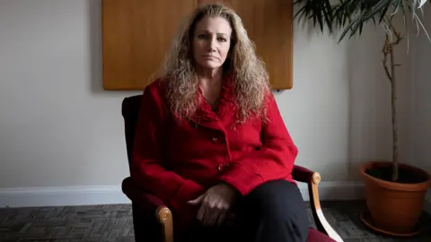 Marlean Ames ints in a chair weraring a red blazer  and black pants with her hair falling over her shoulders