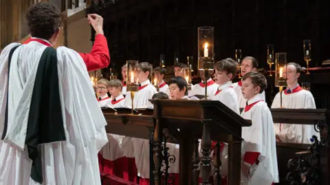 Young choristers from the Choir of King's College, Cambridge, during a final rehearsal at King's College Chapel ahead of the Festival of Nine Lessons and Carols. They are singing and looking at the conductor in front of them, wearing choir robes and surrounded by candles.
