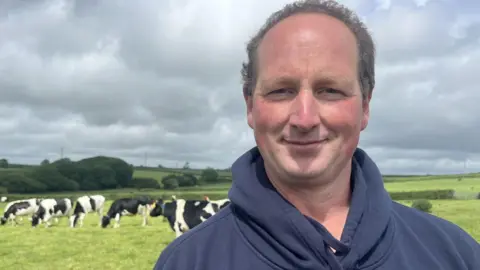 A man in a blue hoodie is looking at the camera, he is balding with brown hair and a suntanned face, He is smiling and behind him are black and white cows grazing in a field and trees in the distance