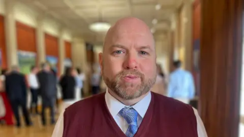 A man with a short grey beard is wearing a white shirt, blue tie and maroon woollen vest. Behind him are groups of people