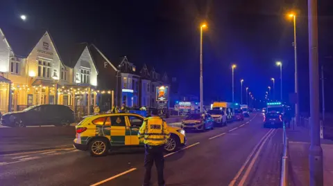 A number of ambulances are parked on a main road at night. On the left of the frame is a large pub. Several police officers are standing by the vehicles. 